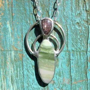 May include: A silver pendant necklace featuring a teardrop-shaped light green stone with horizontal banding, topped by a smaller, faceted, translucent brownish-purple stone. The stones are set in a silver frame with a circular design and chain.