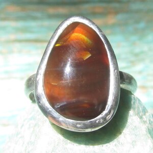 May include: A silver ring featuring a large, teardrop-shaped fire opal gemstone. The stone displays deep reddish-brown tones with internal flashes of orange and yellow light. The metal setting is smooth, polished, and follows the organic shape of the stone.