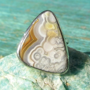 Puede incluir: Anillo de plata con una piedra triangular grande con un patrón único de bandas arremolinadas beige, marrón y blancas. La piedra está engastada en una banda de plata pulida. El anillo se muestra sobre una superficie texturizada de color verde azulado claro.