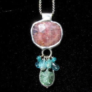 May include: A silver pendant necklace featuring a large, faceted pink gemstone at the top, with a cluster of small blue-green beads and a green gemstone at the bottom. The necklace hangs on a silver chain.