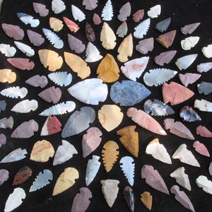May include: A collection of numerous stone arrowheads arranged in a circular pattern. The arrowheads vary in color, including shades of white, gray, tan, brown, red, and black, and feature diverse shapes, sizes, and serrated or smooth edges.