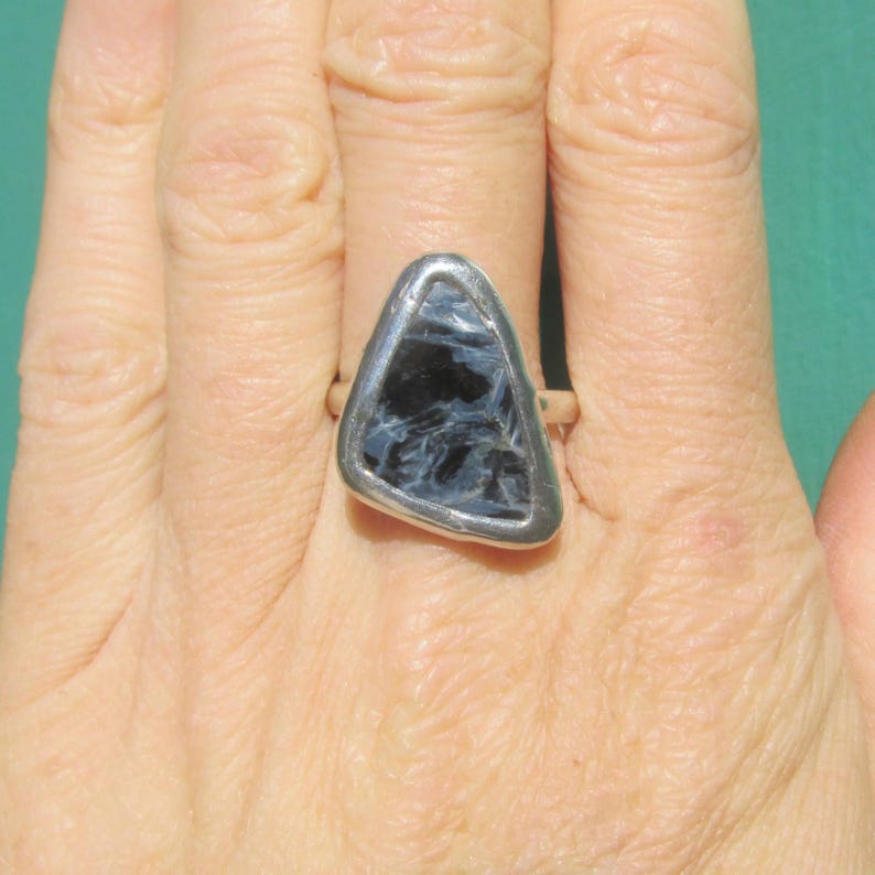 May include: A silver ring featuring a triangular, dark blue gemstone with white veining. The stone is set in a silver bezel, creating a striking contrast. The ring is shown on a hand, showcasing the jewelry's design and color.