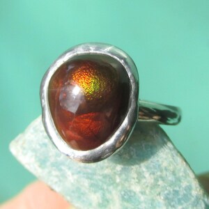 Puede incluir: Anillo de plata con una gema ovalada grande en tonos rojos, marrones y dorados. La gema est&aacute; engastada en una sencilla banda de plata. El anillo se muestra sobre una superficie azul claro. Joya.