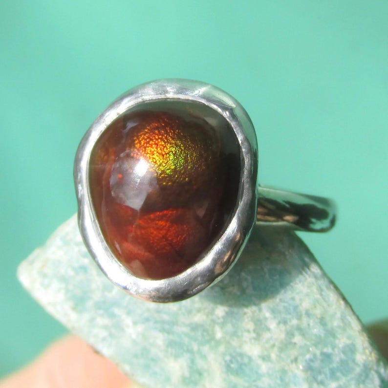 Puede incluir: Anillo de plata con una gema ovalada grande en tonos rojos, marrones y dorados. La gema est&aacute; engastada en una sencilla banda de plata. El anillo se muestra sobre una superficie azul claro. Joya.