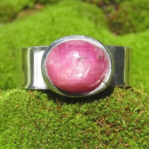 Könnte beinhalten: Ein Silberring mit einem großen, ovalen, im Cabochon-Schliff geschliffenen rosa Edelstein, der in einer glatten Metallfassung sitzt. Die Ringschiene ist breit und poliert und reflektiert das Licht auf ihrer metallischen Oberfläche, während der Stein rosa ist.