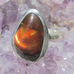 May include: A silver ring featuring a large, teardrop-shaped fire agate gemstone. The stone displays a deep brown base color with a vibrant, glowing orange and red iridescent flash across its polished, smooth surface, set in a simple metal band.
