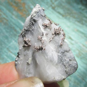 May include: A raw mineral specimen featuring a translucent white quartz base with embedded metallic copper-colored crystalline formations and patches of dark grey matrix material distributed across the jagged, uneven surface of the stone.