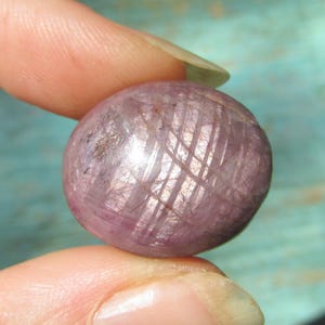 May include: A polished, oval-shaped cabochon gemstone with a translucent, dusty pink and mauve hue. The surface displays a distinct, intersecting grid-like pattern of fine white inclusions that create a subtle, light-reflecting star effect.