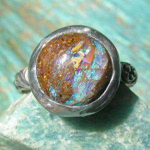 May include: A silver ring featuring a circular, polished stone inlay. The stone displays a textured, earthy brown surface on one side, transitioning into vibrant, iridescent flashes of blue, purple, and pink across the remaining surface area.