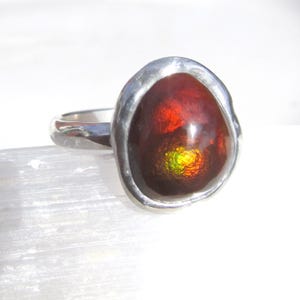 Puede incluir: Anillo de plata con una piedra preciosa grande y ovalada. La piedra tiene un aspecto ardiente con tonos rojos, naranjas y amarillos. El anillo est&aacute; engastado en una banda de plata lisa y pulida. El anillo se muestra sobre una superficie blanca.