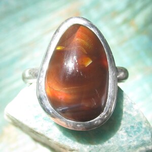 May include: A silver ring featuring a smooth, teardrop-shaped fire agate gemstone. The stone displays deep reddish-brown tones with internal flashes of orange and yellow light, securely set within a polished, solid silver bezel and band.