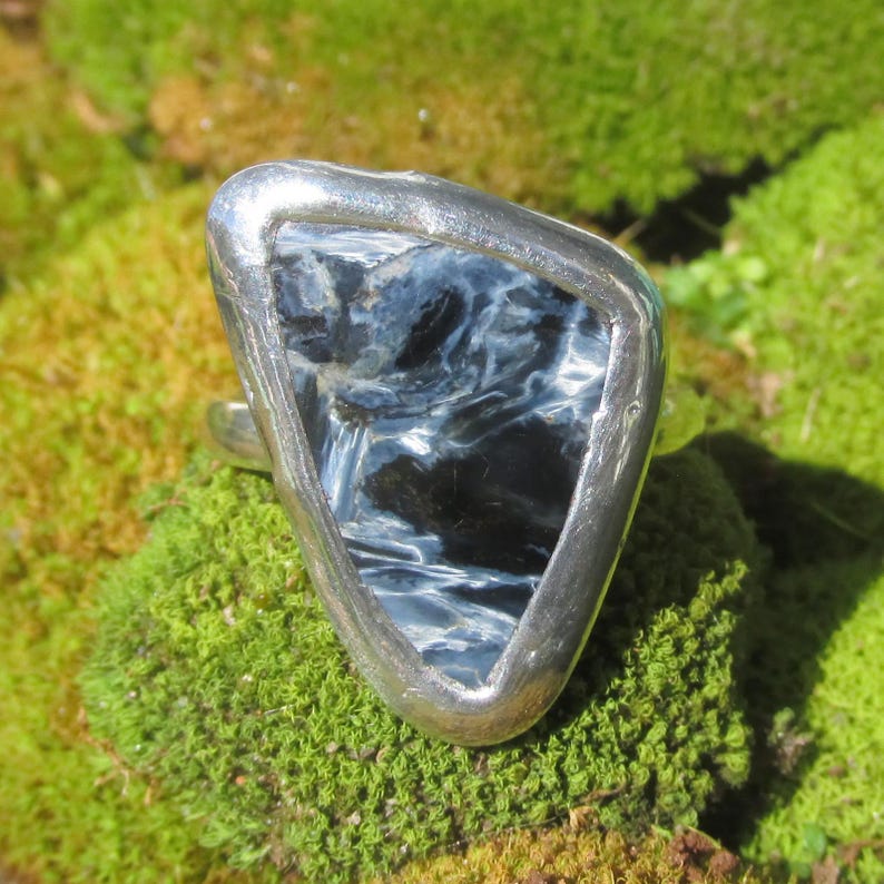 May include: A silver ring featuring a triangular, polished gemstone with a swirling pattern of blue and black. The ring is set against a backdrop of green moss, highlighting the natural colors and textures of the jewelry.