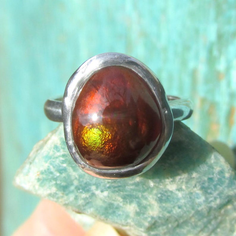 Puede incluir: Un anillo de plata con una piedra preciosa ovalada grande. La piedra preciosa tiene una exhibici&oacute;n ardiente de colores rojo, naranja y verde. El anillo est&aacute; engastado en una banda de plata pulida. Las dimensiones de la piedra preciosa no son visibles.
