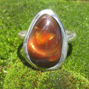May include: A silver ring featuring a large, teardrop-shaped fire agate gemstone. The stone is deep brown with internal flashes of bright orange and yellow light. The metal band is smooth, polished, and surrounds the stone in a simple setting.