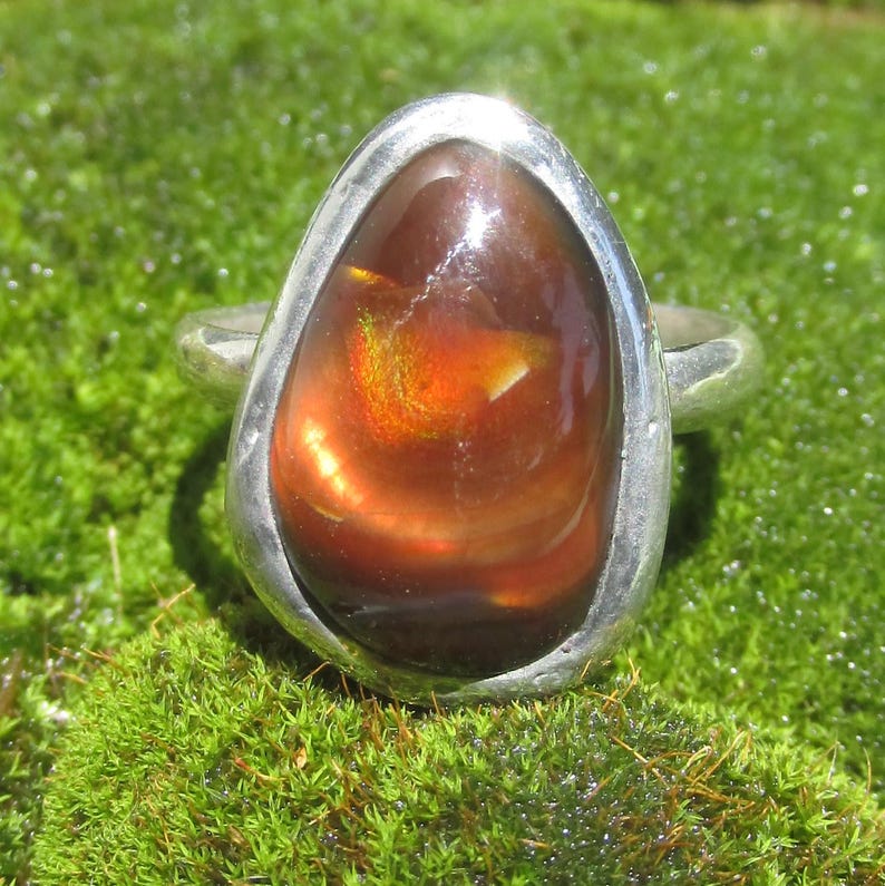May include: A silver ring featuring a large, teardrop-shaped fire agate gemstone. The stone is deep brown with internal flashes of bright orange and yellow light. The metal band is smooth, polished, and surrounds the stone in a simple setting.