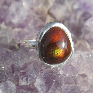 Puede incluir: Un anillo de plata con una gran gema ovalada. La gema tiene una apariencia ardiente e iridiscente, con tonos rojos, naranjas y verdes. El anillo est&aacute; engastado sobre un fondo de cristales morados.