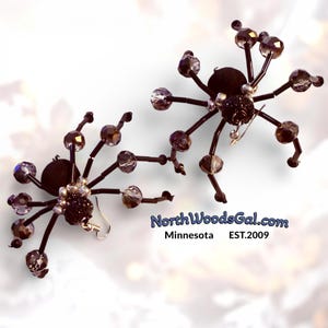 Puede incluir: Pendientes de araña con cuerpos de terciopelo negro, patas de alambre negro y cuentas de cristal facetadas. Los pendientes tienen ganchos plateados. El texto "NorthWoodsGal.com Minnesota EST.2009" es visible.