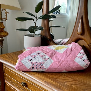 Peut inclure: Une courtepointe rose et blanche en patchwork avec un motif floral pliée sur un commode en bois avec un tiroir.