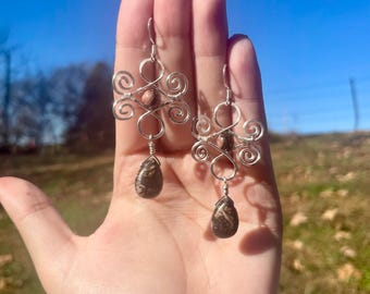 May include: Silver wire earrings featuring a swirling design and teardrop-shaped brown and black stones. The earrings are shown against a blue sky background.