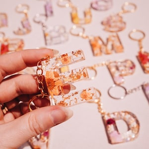May include: A clear resin letter "E" keyring, filled with dried orange flowers and gold flakes, held in a hand. Other resin letter keyrings with flowers and gold accents are in the background, attached to gold keyrings.