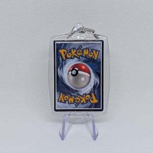 May include: A rectangular clear acrylic keychain case containing a standard blue and yellow Pok&eacute;mon trading card back design. The card features a central red and white Pok&eacute; Ball graphic surrounded by a swirling blue vortex pattern and the Pok&eacute;mon logo.