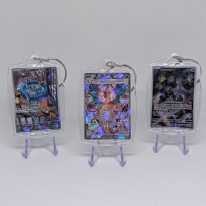 May include: Three rectangular clear acrylic keychains, each housing a holographic trading card. The left keychain features a blue character, the center a multi-colored winged creature, and the right a dark purple figure. Each is displayed on a small clear stand.
