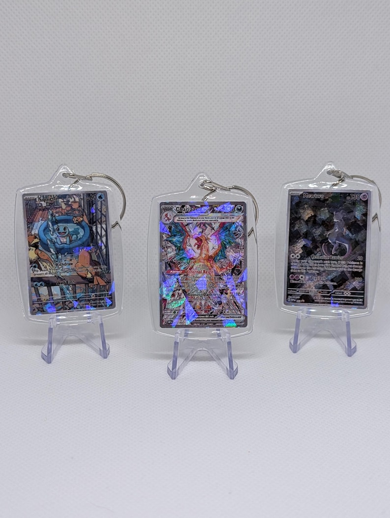 May include: Three rectangular clear acrylic keychains, each housing a holographic trading card. The left keychain features a blue character, the center a multi-colored winged creature, and the right a dark purple figure. Each is displayed on a small clear stand.
