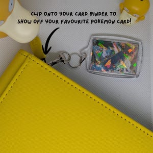 May include: A clear acrylic card holder keychain attached to a yellow binder with a metal clasp. The holder displays a holographic trading card. Text reads: CLIP ONTO YOUR CARD BINDER TO SHOW OFF YOUR FAVOURITE POKEMON CARD! Two yellow Psyduck figures are visible.