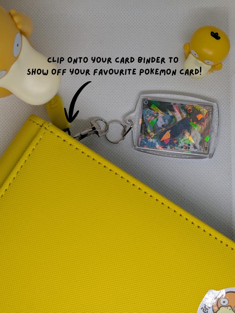 May include: A clear acrylic card holder keychain attached to a yellow binder with a metal clasp. The holder displays a holographic trading card. Text reads: CLIP ONTO YOUR CARD BINDER TO SHOW OFF YOUR FAVOURITE POKEMON CARD! Two yellow Psyduck figures are visible.