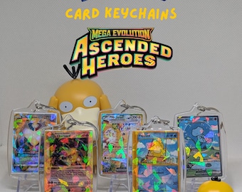 May include: Mini Pokémon card keychains featuring holographic trading cards encased in clear acrylic protectors. The display includes text reading MINI POKÉMON CARD KEYCHAINS and MEGA EVOLUTION ASCENDED HEROES, alongside two yellow Psyduck figures.