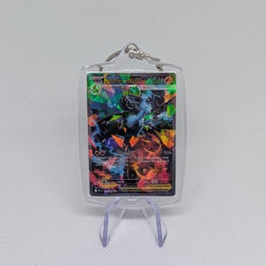May include: A clear acrylic keychain holder containing a holographic Mega Charizard X EX trading card. The card features a dark dragon illustration with colorful geometric foil patterns, text reading Mega Charizard X EX, and a 360 HP value.