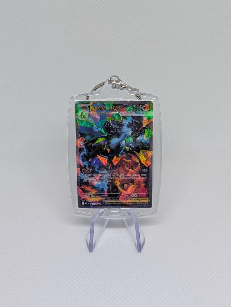 May include: A clear acrylic keychain holder containing a holographic Mega Charizard X EX trading card. The card features a dark dragon illustration with colorful geometric foil patterns, text reading Mega Charizard X EX, and a 360 HP value.