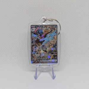 May include: A clear acrylic keychain containing a Greninja ex Pok&eacute;mon trading card with a holographic finish. The card features the character Greninja, the text Greninja ex, 300 HP, and the attacks Stealth Shuriken and Torrential Slash.