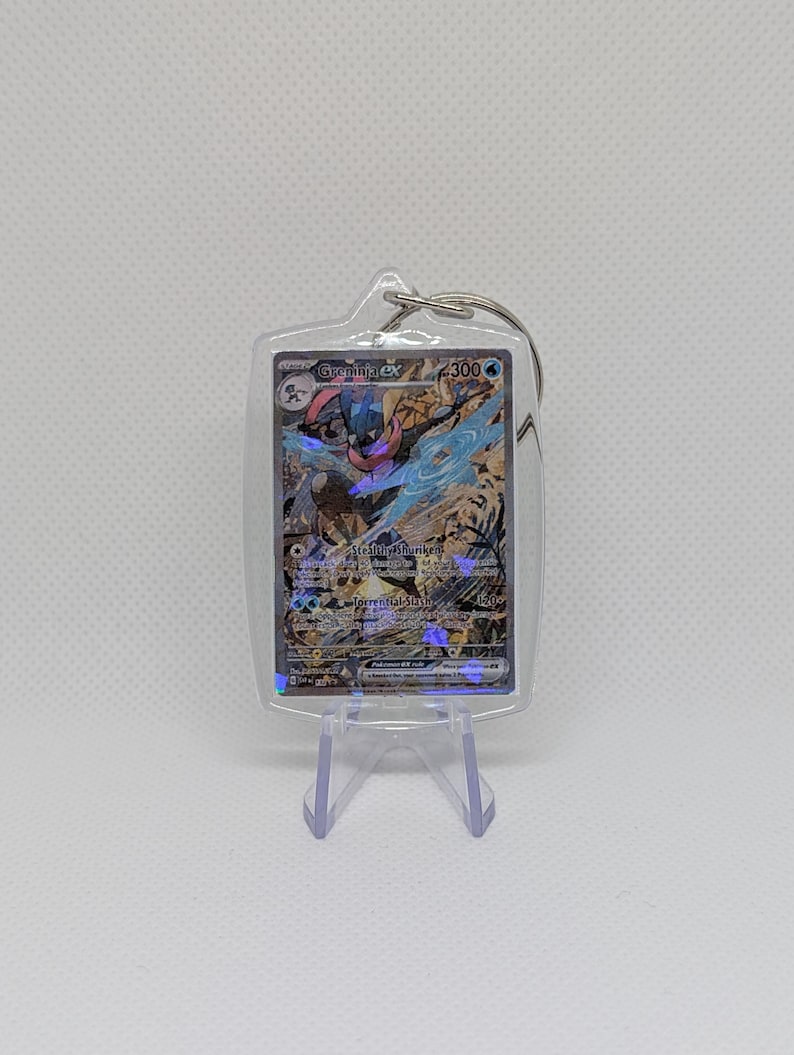 May include: A clear acrylic keychain containing a Greninja ex Pok&eacute;mon trading card with a holographic finish. The card features the character Greninja, the text Greninja ex, 300 HP, and the attacks Stealth Shuriken and Torrential Slash.