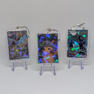 May include: Three rectangular clear acrylic keychains, each housing a holographic Pok&eacute;mon trading card. The cards feature Greninja ex, Detective Pikachu, and Umbreon VMAX. Each keychain includes a metal ring and rests on a small clear stand.