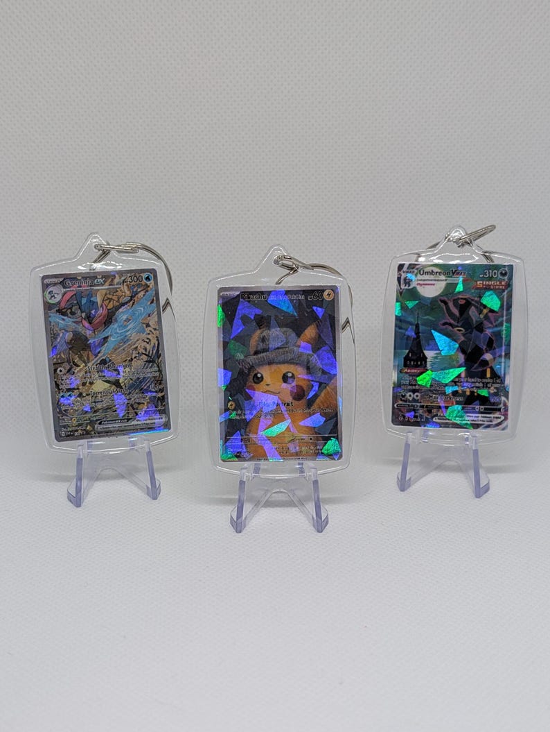 May include: Three rectangular clear acrylic keychains, each housing a holographic Pok&eacute;mon trading card. The cards feature Greninja ex, Detective Pikachu, and Umbreon VMAX. Each keychain includes a metal ring and rests on a small clear stand.