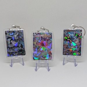 May include: Three rectangular clear acrylic keychains, each housing a holographic trading card. The cards feature colorful illustrations of fantasy creatures with visible text and numbers, displayed upright on small clear plastic stands.