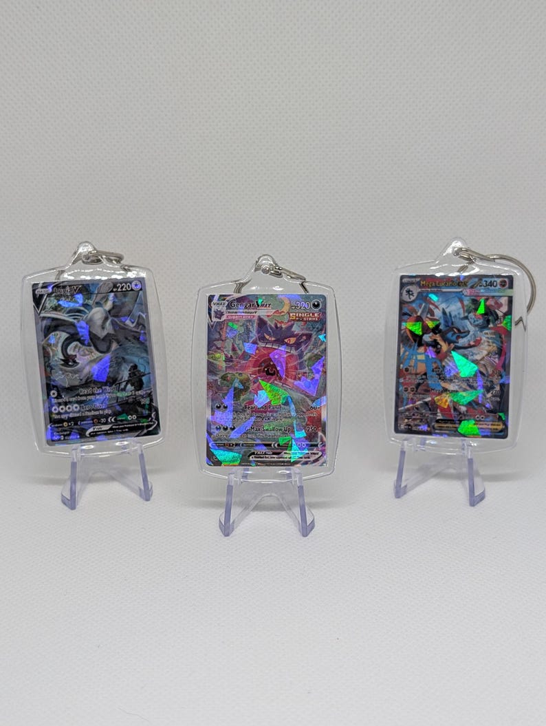 May include: Three rectangular clear acrylic keychains, each housing a holographic trading card. The cards feature colorful illustrations of fantasy creatures with visible text and numbers, displayed upright on small clear plastic stands.
