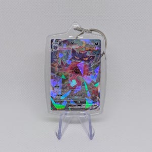 May include: A clear acrylic keychain case containing a Gengar VMAX trading card. The card features a purple ghost character with a wide mouth, 320 HP, and Single Strike text. The surface has a holographic finish with geometric shards.