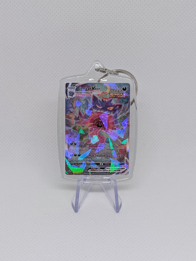 May include: A clear acrylic keychain case containing a Gengar VMAX trading card. The card features a purple ghost character with a wide mouth, 320 HP, and Single Strike text. The surface has a holographic finish with geometric shards.