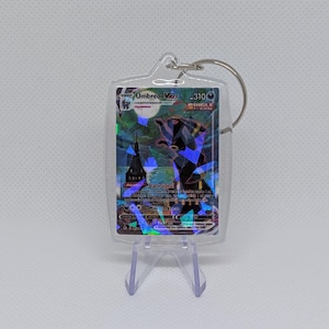 May include: A clear acrylic keychain case containing a holographic Umbreon VMAX trading card. The card features a dark creature, a full moon, and a building silhouette with the text Umbreon VMAX, 310 HP, and Single Strike. A metal ring is attached.