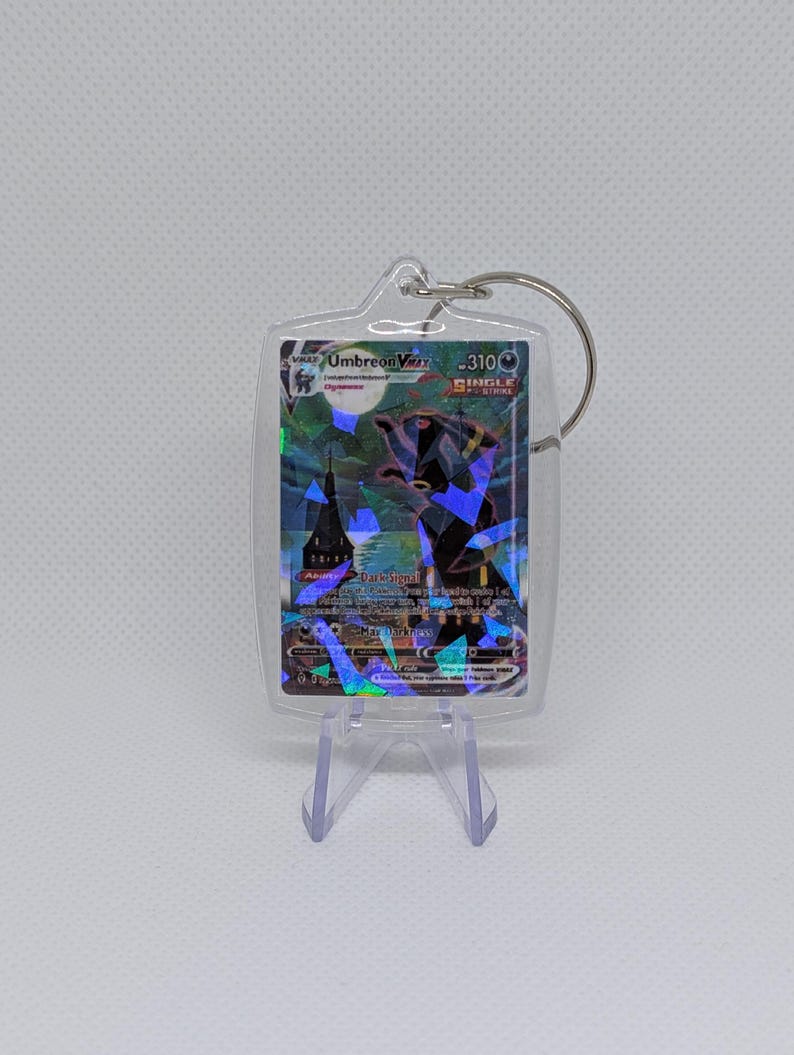 May include: A clear acrylic keychain case containing a holographic Umbreon VMAX trading card. The card features a dark creature, a full moon, and a building silhouette with the text Umbreon VMAX, 310 HP, and Single Strike. A metal ring is attached.