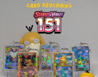 May include: A collection of five miniature Pokémon trading cards encased in clear acrylic keychain holders, featuring holographic foil patterns. The text MINI POKÉMON CARD KEYCHAINS SCARLET & VIOLET 151 is displayed above. Two yellow Psyduck figurines are present.