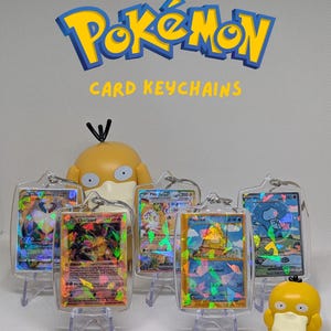 May include: Five mini Pok&eacute;mon card keychains in clear protective cases, each featuring a holographic trading card. The text MINI POK&Eacute;MON CARD KEYCHAINS is displayed above. Two yellow Psyduck figurines are positioned among the keychains.