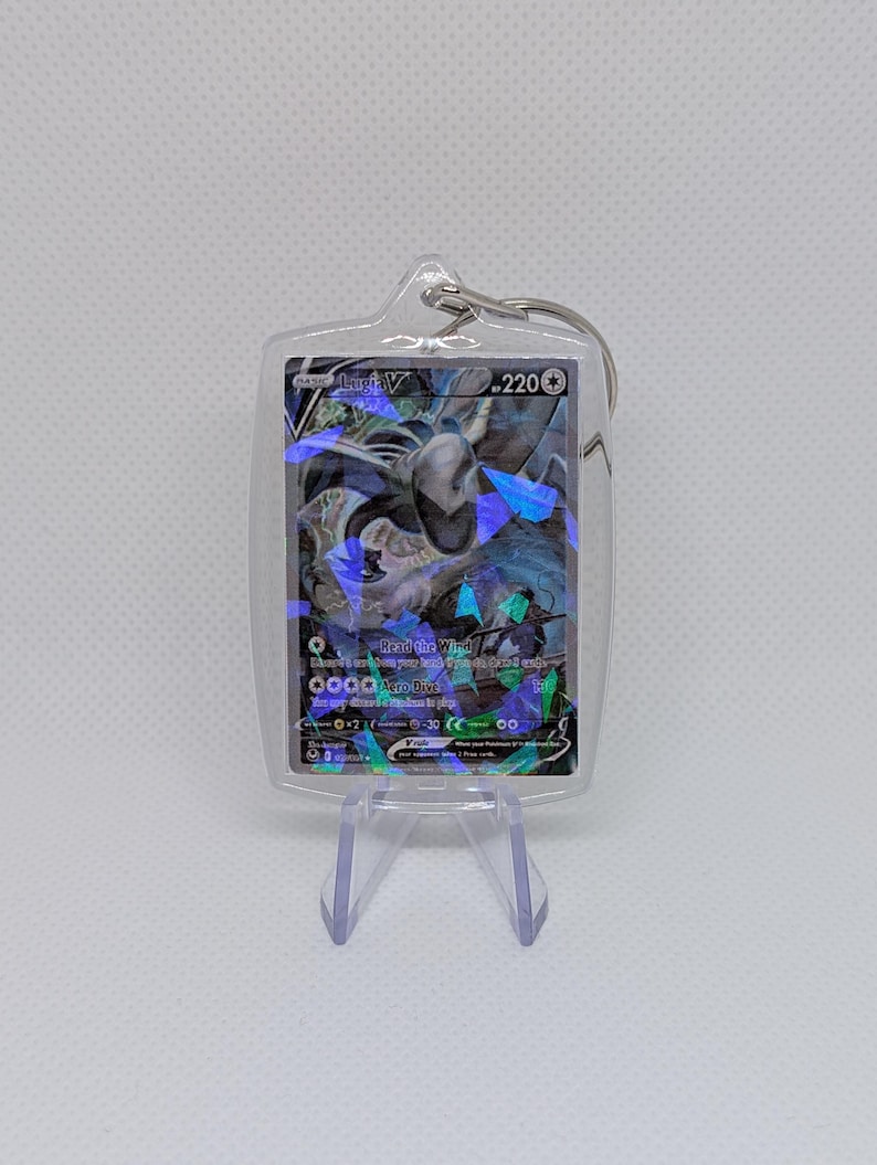 May include: A clear acrylic keychain containing a Lugia V trading card with 220 HP. The card features a holographic finish with blue and purple geometric shards, displaying the text Lugia V, 220, Read the Wind, and Aero Dive.