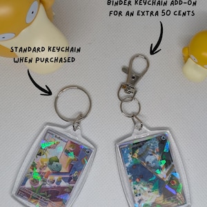 May include: Two rectangular acrylic keychains featuring holographic trading cards. One has a standard metal split ring, and the other has a metal lobster clasp. Text reads: STANDARD KEYCHAIN WHEN PURCHASED and BINDER KEYCHAIN ADD-ON FOR AN EXTRA 50 CENTS.