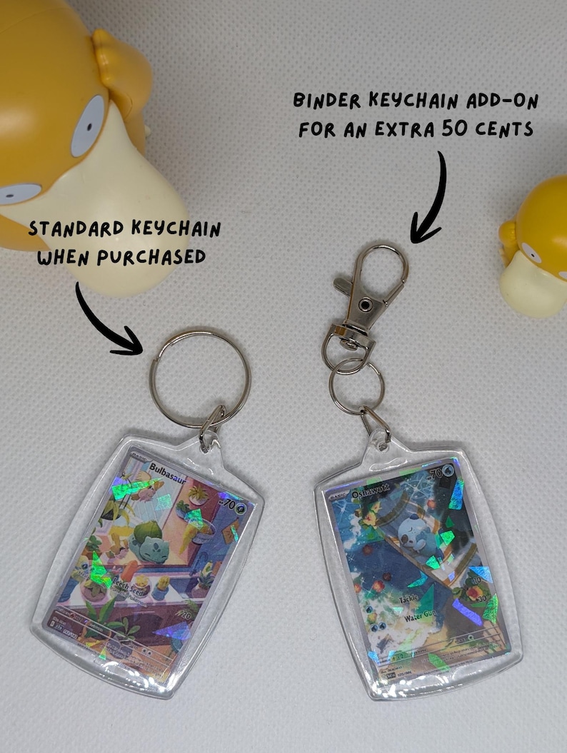 May include: Two rectangular acrylic keychains featuring holographic trading cards. One has a standard metal split ring, and the other has a metal lobster clasp. Text reads: STANDARD KEYCHAIN WHEN PURCHASED and BINDER KEYCHAIN ADD-ON FOR AN EXTRA 50 CENTS.