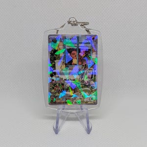May include: A clear acrylic keychain featuring a Charmander trading card with a holographic finish. The card displays the name Charmander, 60 HP, and the move Heat Tackle with 30 damage. The keychain is held in a transparent plastic case.