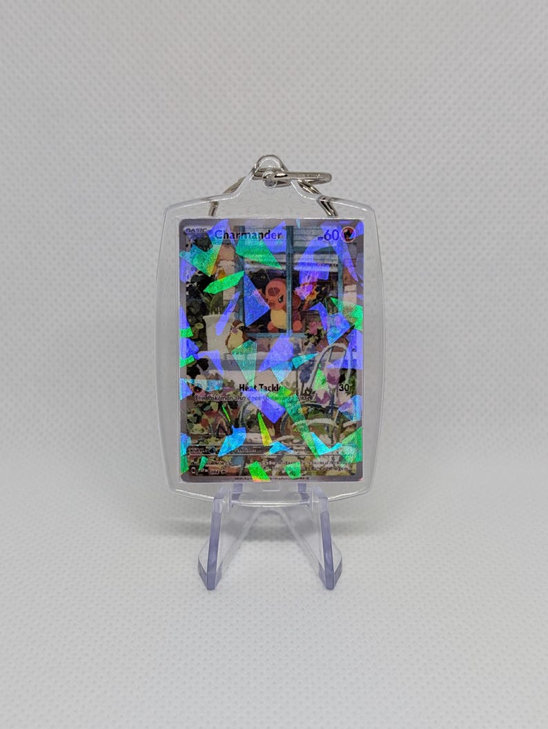 May include: A clear acrylic keychain featuring a Charmander trading card with a holographic finish. The card displays the name Charmander, 60 HP, and the move Heat Tackle with 30 damage. The keychain is held in a transparent plastic case.