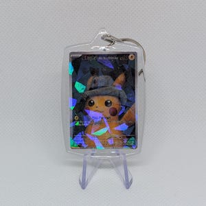 May include: A clear acrylic keychain containing a holographic trading card featuring a yellow Pikachu wearing a grey hat. The card displays the text Pikachu, Pika-Portrait, and 60 HP. The keychain has a metal ring at the top.